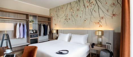 A hotel room with a large bed, a desk with a chair, a wardrobe, and a wall-mounted lamp.