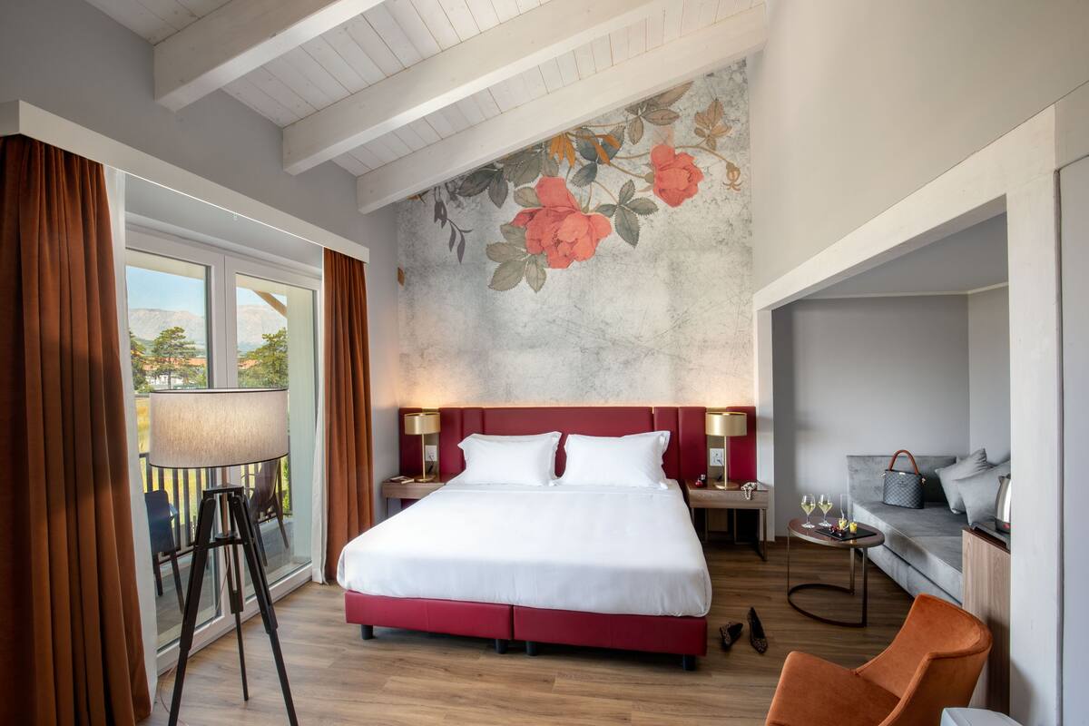 Quarto de hotel com uma cama grande, varanda, sofá e uma pequena mesa com bebidas.