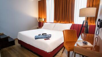 Una camera d'albergo con un letto, una scrivania, una sedia, una lampada e una finestra con tende.