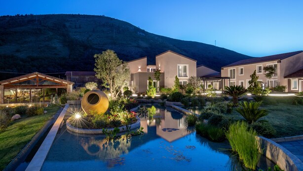 A modern hotel with a swimming pool, landscaped gardens, and a mountainous backdrop.