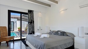 A modern bedroom with a large bed, a chair, a nightstand, and a view of an outdoor pool area. - Villa Marcelino with Private Pool and free A/C near to Port de Pollensa (Puerto Pollensa)