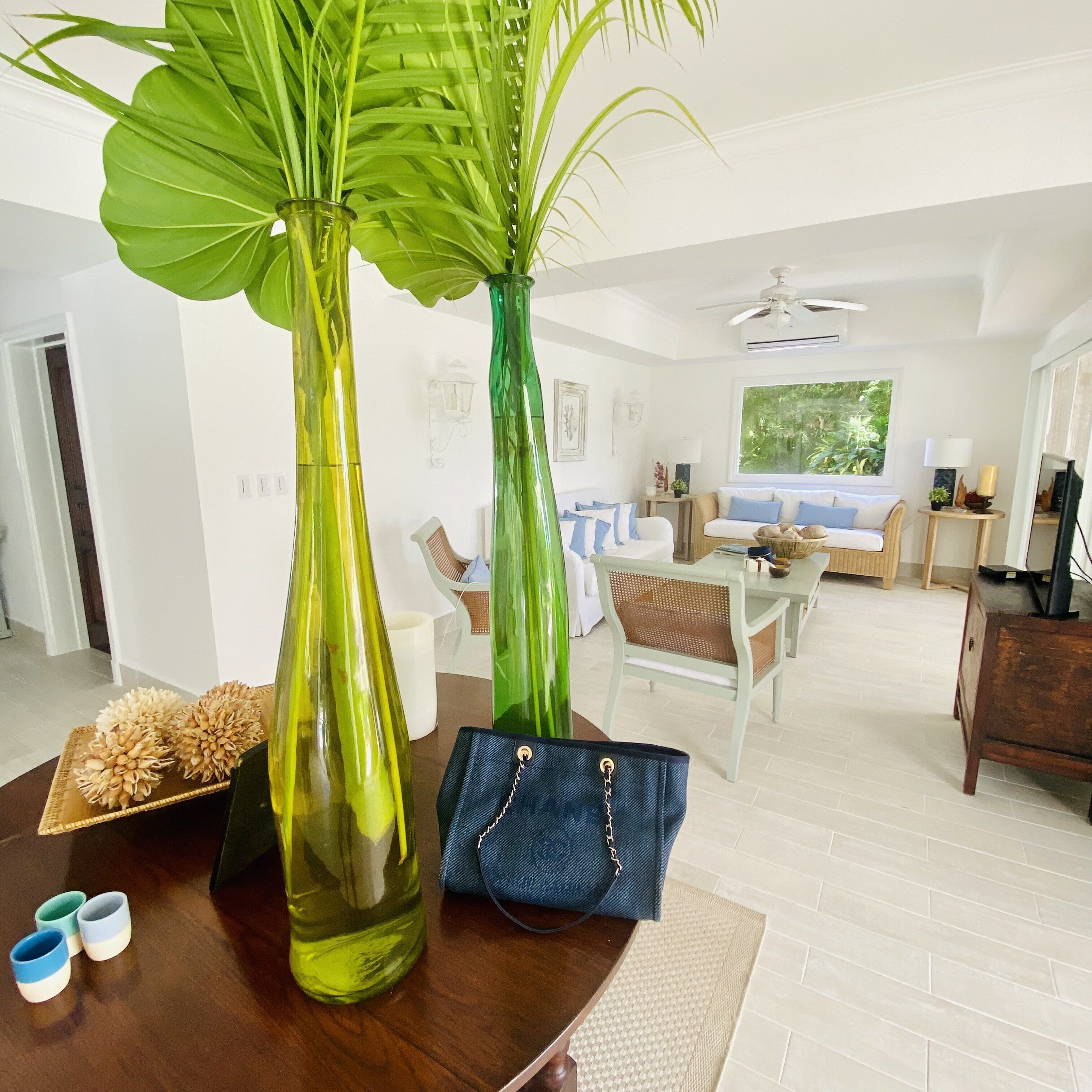 Een woonkamer met een eethoek, met een grote groene plant in een vaas, een blauwe handtas en een houten tafel met decoratieve objecten.