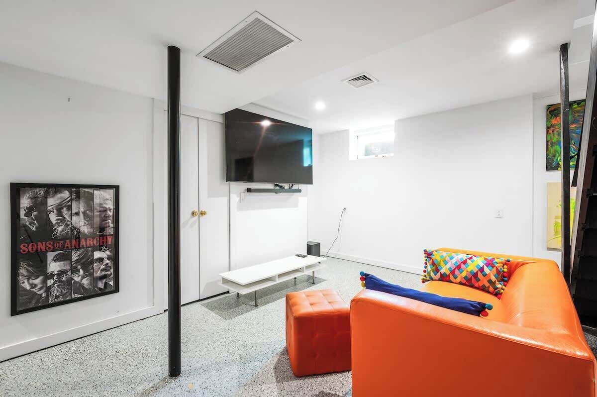 A modern living room with a flat-screen TV, a bench, and an orange sofa with colorful pillows.