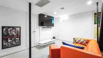 A modern living room with a flat-screen TV, a bench, and an orange sofa with colorful pillows.