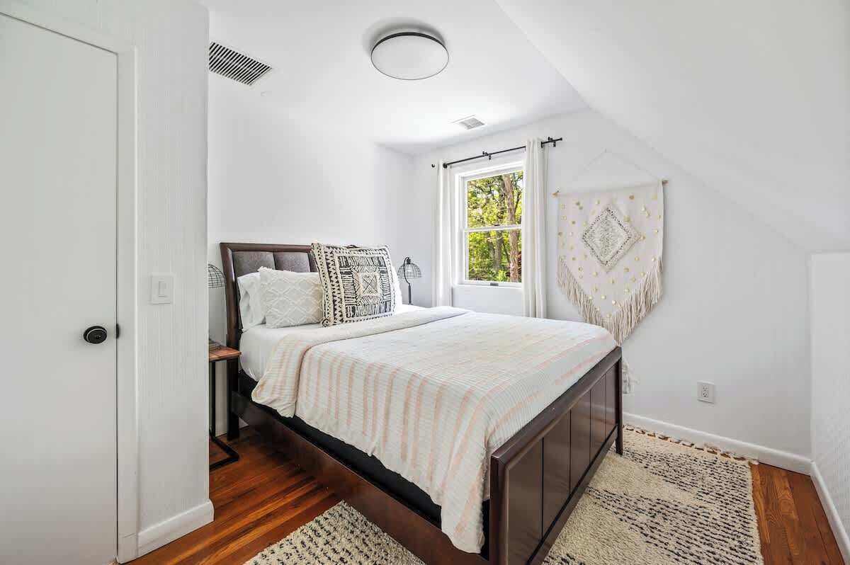 Une chambre à coucher avec un lit, une fenêtre avec des rideaux, une porte et une bouche d’aération au plafond.