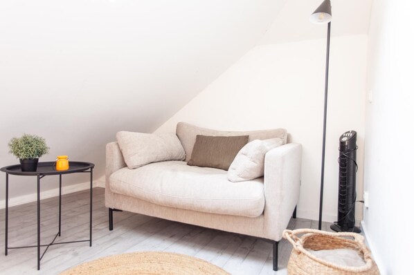 Una habitación acogedora con un sofá color beige, una pequeña mesa redonda con una planta en maceta, una lámpara de pie y un calefactor.