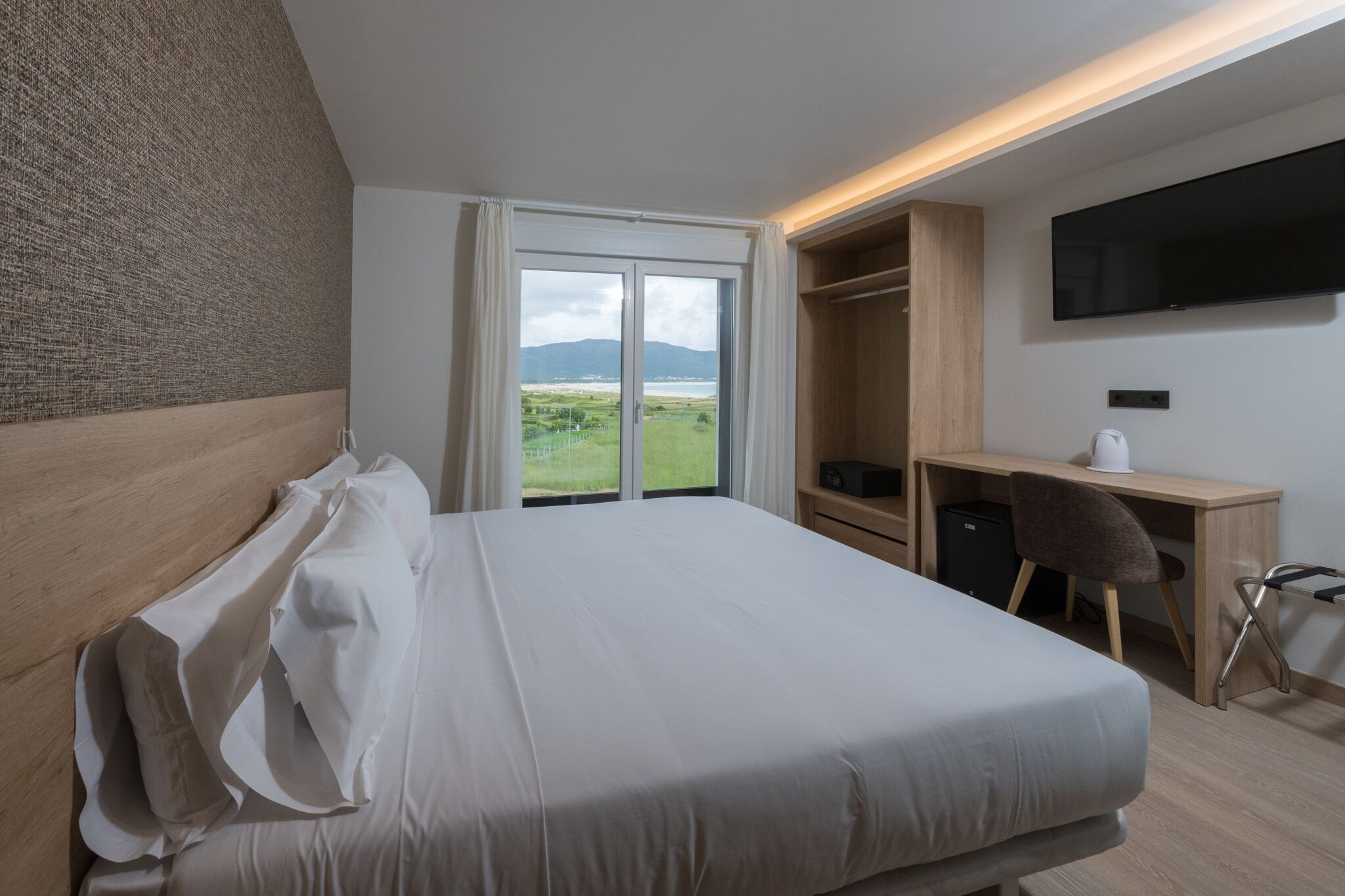 Et hotelværelse med en stor seng, et skrivebord, en stol, et fjernsyn og udsigt over et grønt landskab gennem en skydedør.