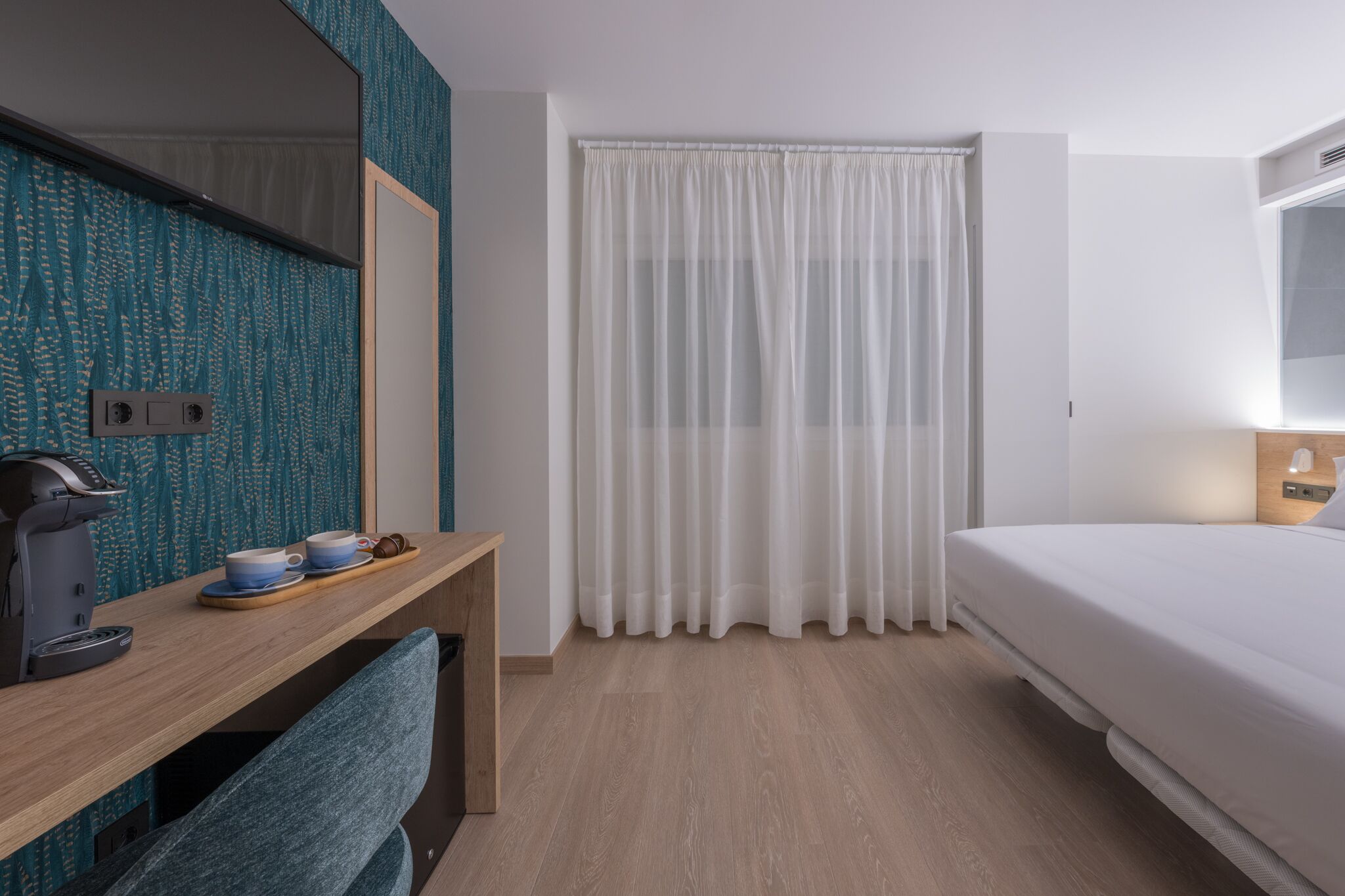Quarto de hotel com secretária de madeira, máquina de café, cama e janela com cortinas brancas.