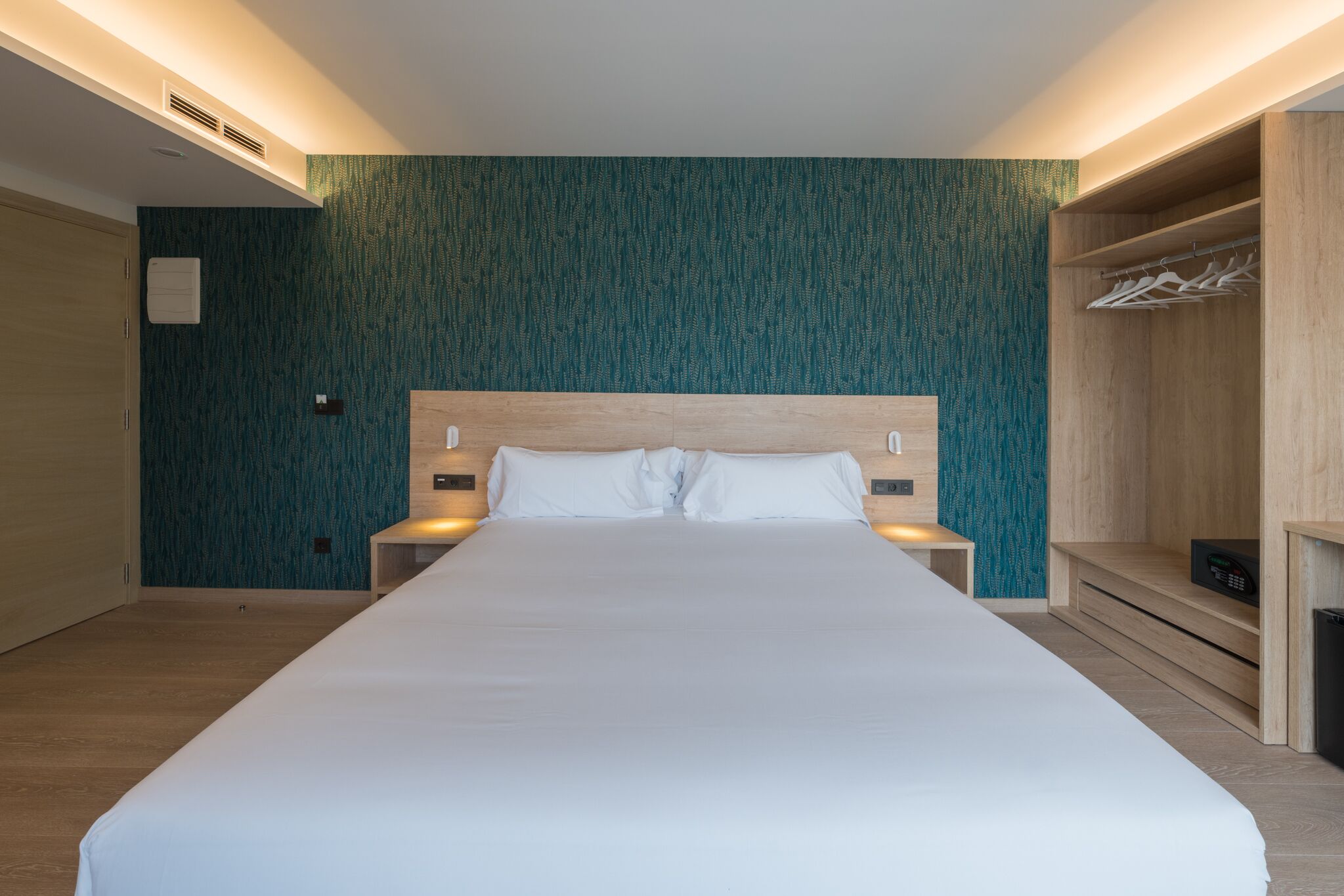 Une chambre d’hôtel moderne avec un grand lit, une tête de lit en bois, un mur orné d’un motif turquoise et une armoire intégrée avec espace pour suspendre les vêtements.