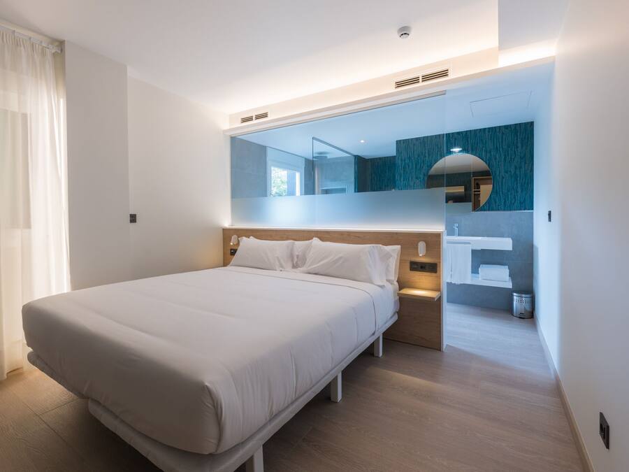 A modern hotel room with a large bed, a bedside table, a mirror, and a view into the bathroom.