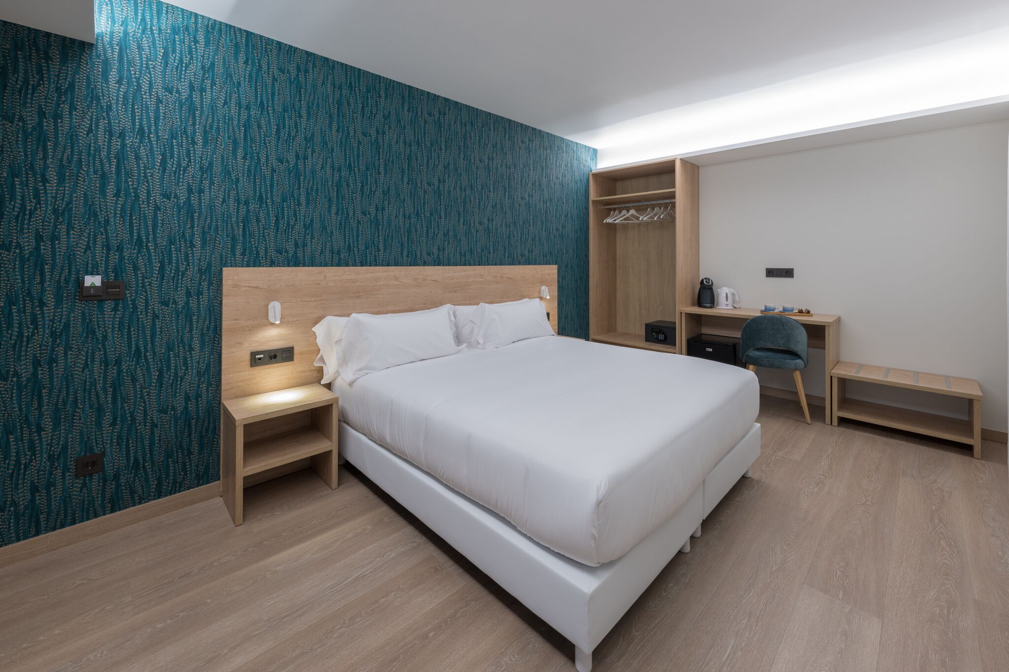 A modern hotel room with a large bed, a wooden nightstand, a desk with a chair, and a wardrobe.