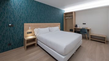 A modern hotel room with a large bed, a wooden nightstand, a desk with a chair, and a wardrobe.