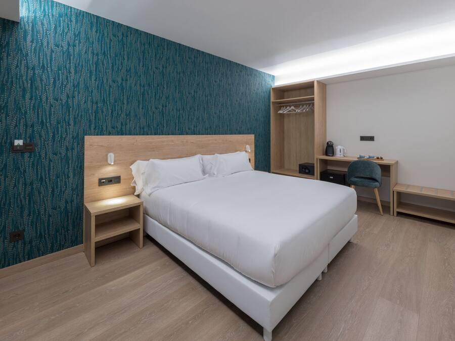 A modern hotel room with a large bed, a wooden nightstand, a desk with a chair, and a wardrobe.
