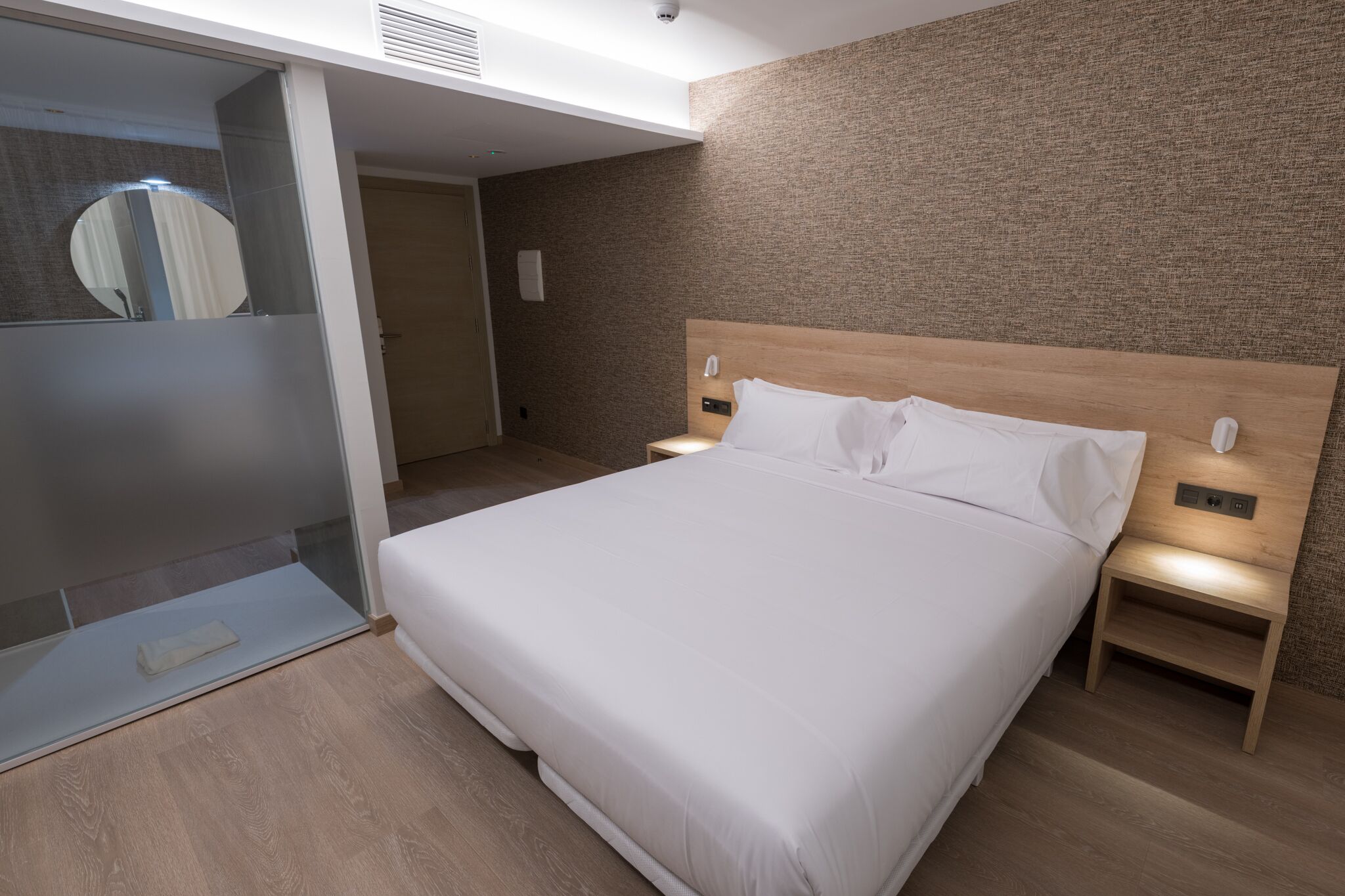 Une chambre d’hôtel moderne avec un grand lit, une tête de lit en bois, des lampes de chevet et une cabine de douche en verre.