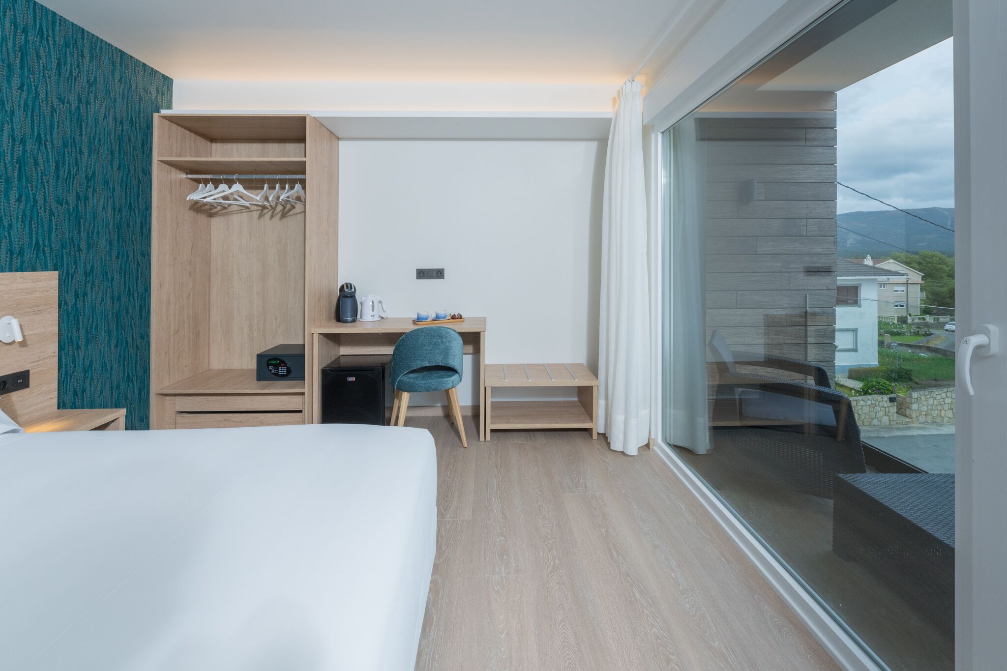 A modern hotel room with a bed, a desk with a chair, a wardrobe, and a balcony with a view of the outside.