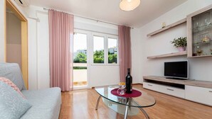 Un salon moderne avec une table basse en verre, un canapé gris, un téléviseur à écran plat posé sur une étagère en bois et une porte-fenêtre coulissante donnant sur un balcon.