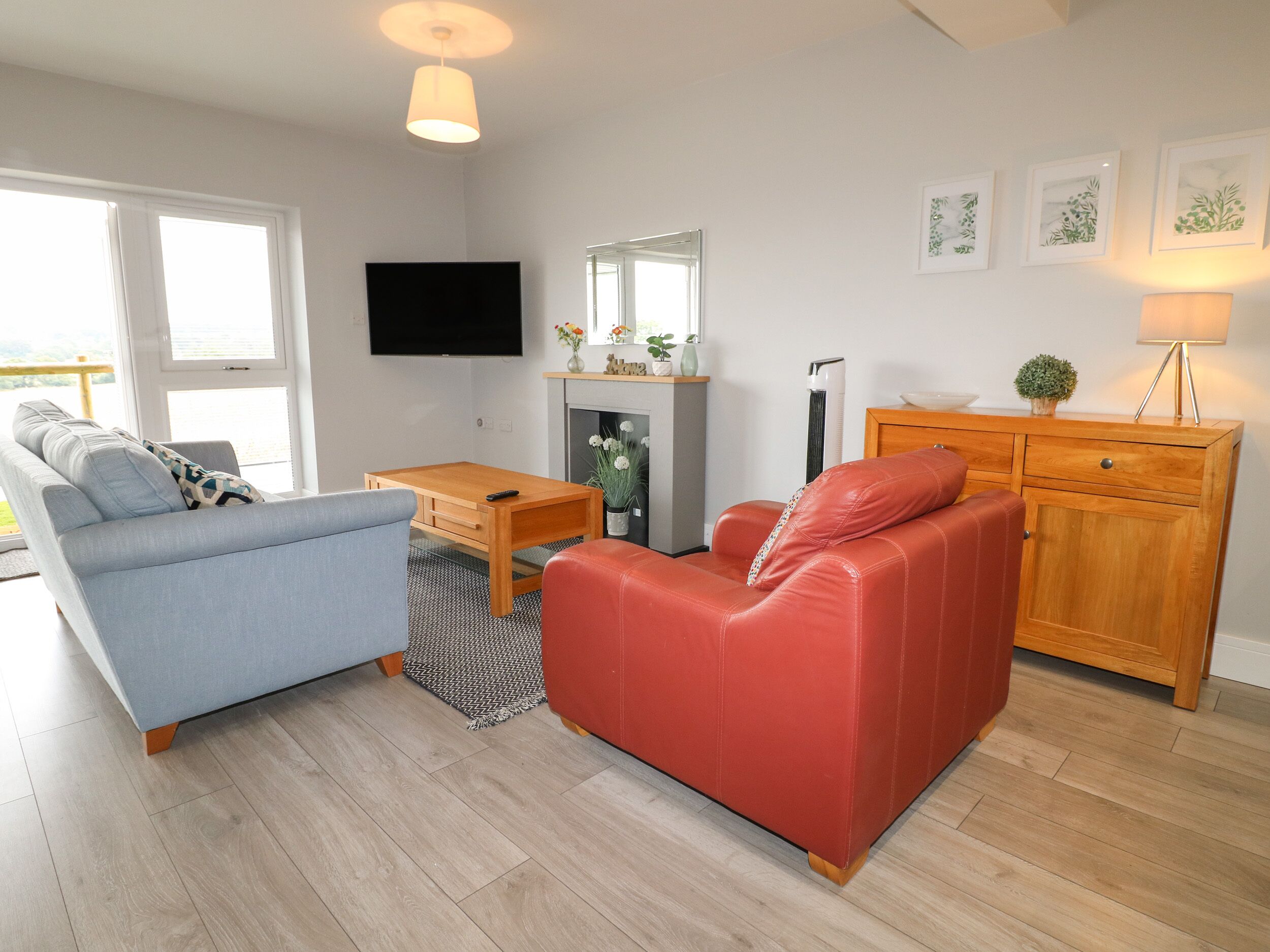 A living room with a sofa, a red armchair, a wooden coffee table, a flat-screen TV mounted on the wall, a fireplace, and a wooden cabinet.