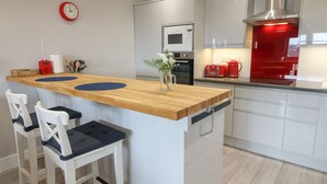 Une cuisine moderne avec un îlot central en bois, des tabourets de bar blancs, une horloge rouge et des appareils électroménagers en acier inoxydable.