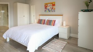 Une chambre à coucher moderne avec un grand lit, des tables de chevet blanches, une table de chevet avec une lampe, une peinture abstraite colorée au mur et une armoire blanche.