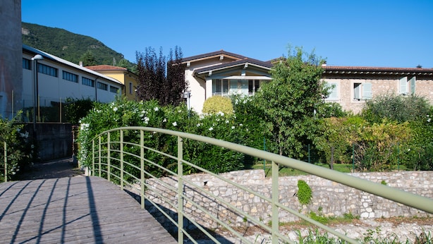 Un chemin de pierres menant à un bâtiment doté d’un balcon, entouré de verdure et surplombé d’un ciel bleu clair.
