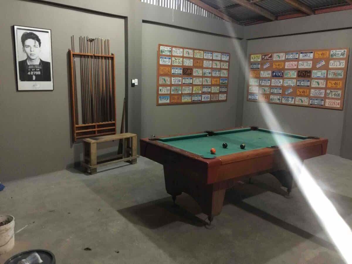 Una mesa de billar con un rayo de luz en una habitación de piso de concreto. Hay tacos y una foto enmarcada en la pared.