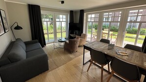 Uma sala de estar moderna com um sofá, uma mesa de jantar com cadeiras, uma TV e janelas amplas com vista para o exterior.
