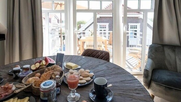 A dining table with breakfast items, a sofa, and a view of buildings through large windows. - Wattkieker in the Lindenhof - centrally located with a classic design (Wangerooge)