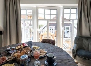 A dining table with breakfast items, a sofa, and a view of buildings through large windows.