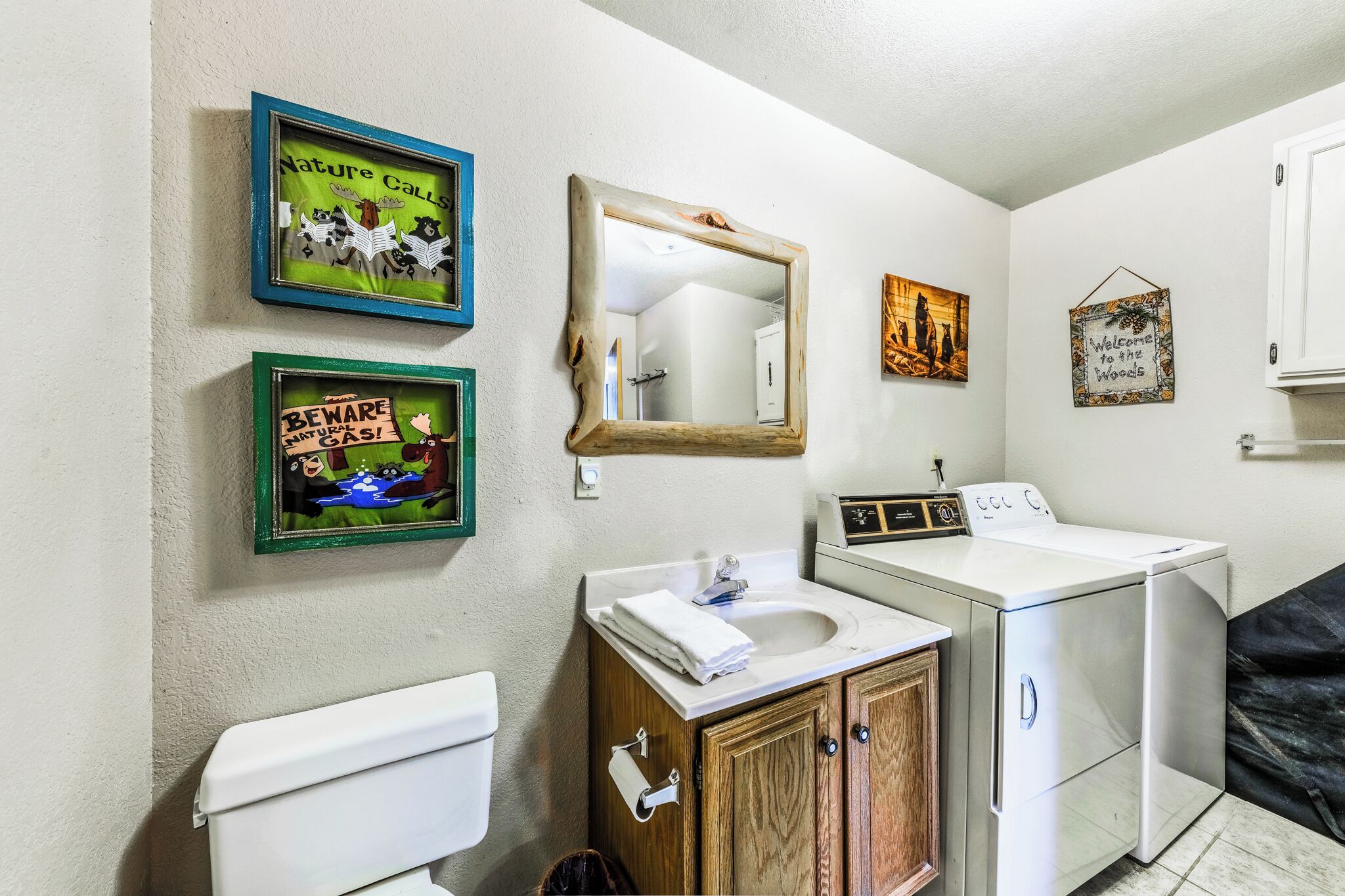 A bathroom with a toilet, sink, mirror, and washing machine. There are framed pictures on the wall.