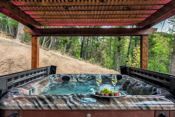 Relax in a serene hot tub under a shaded pergola with a view of the surrounding forest.