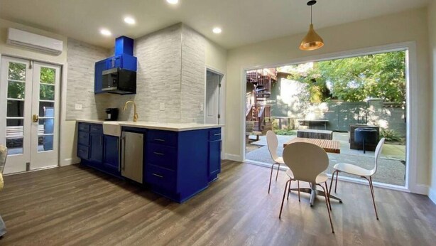 Uma cozinha moderna com armários azuis, painel de fundo branco e uma ilha grande. Há uma área de jantar com duas cadeiras e uma mesa, e uma porta de vidro deslizante que dá para um quintal com móveis de exterior.