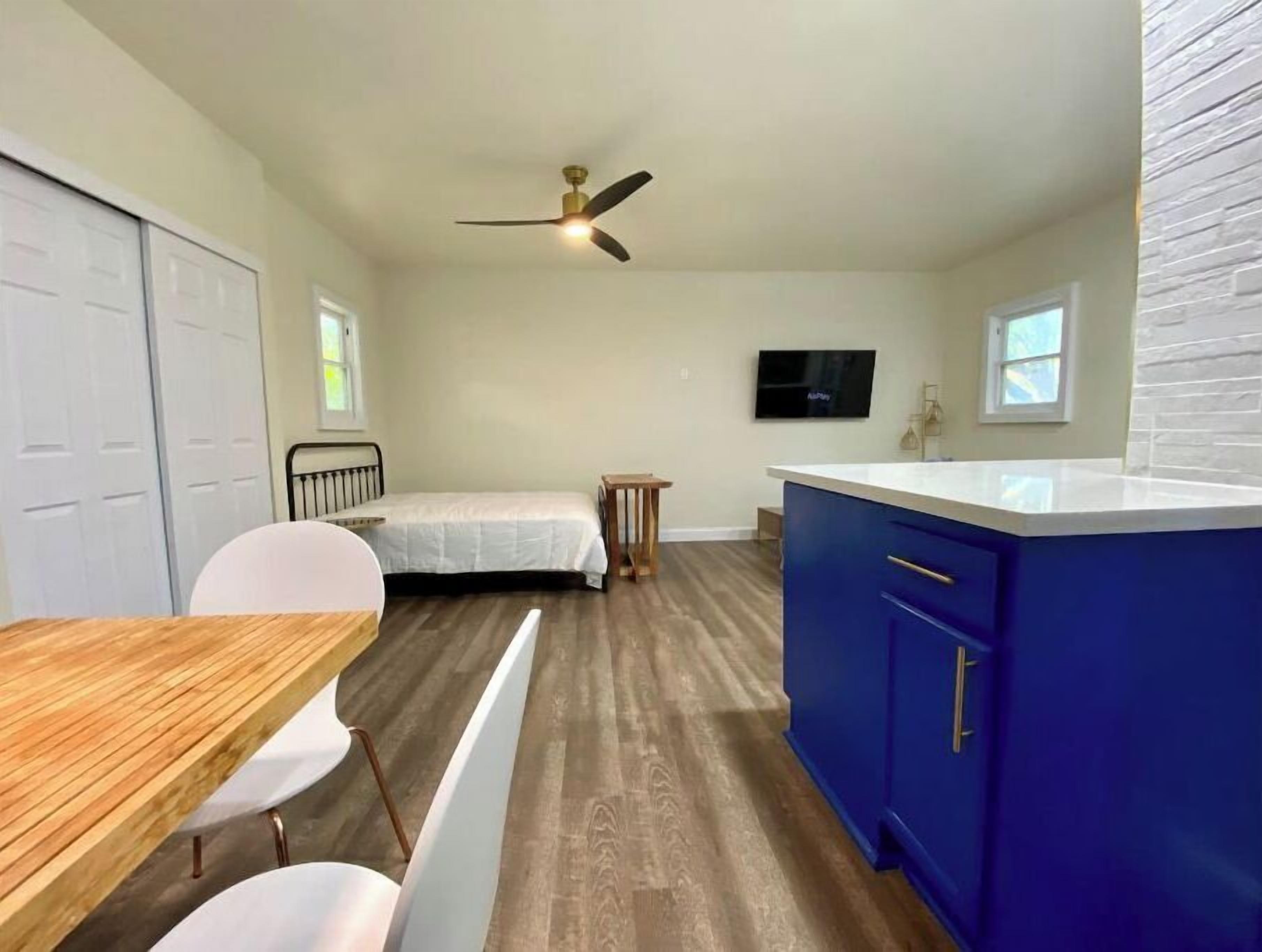 Une chambre à coucher de taille modeste, comprenant un lit, un bureau et une chaise. On peut voir un ventilateur de plafond, et un téléviseur est fixé au mur.