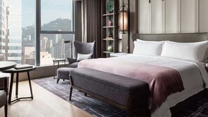 A modern hotel room with a large bed, a seating area with a chair and table, a bookshelf, and a view of the city. - HUALUXE Qingdao Licang by IHG (Qingdao)