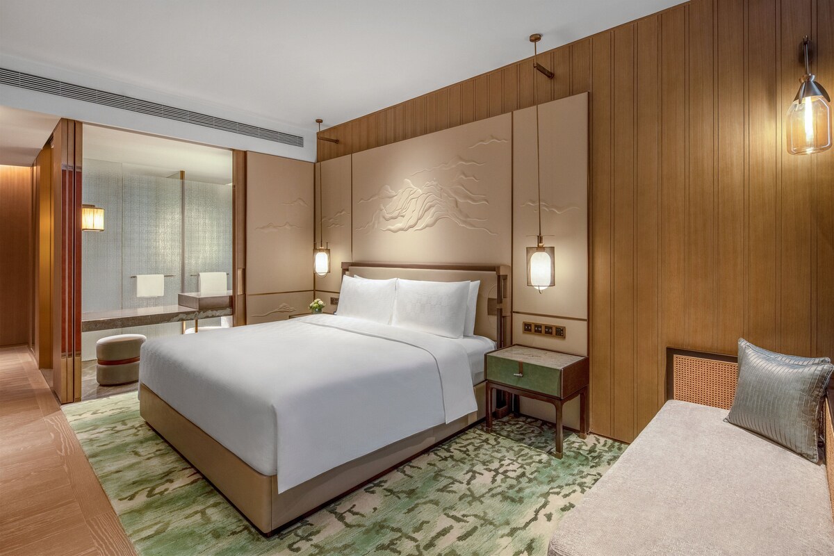 A hotel room with a large bed, a bedside table, a nightstand, a mirror, and a wall with a decorative panel.