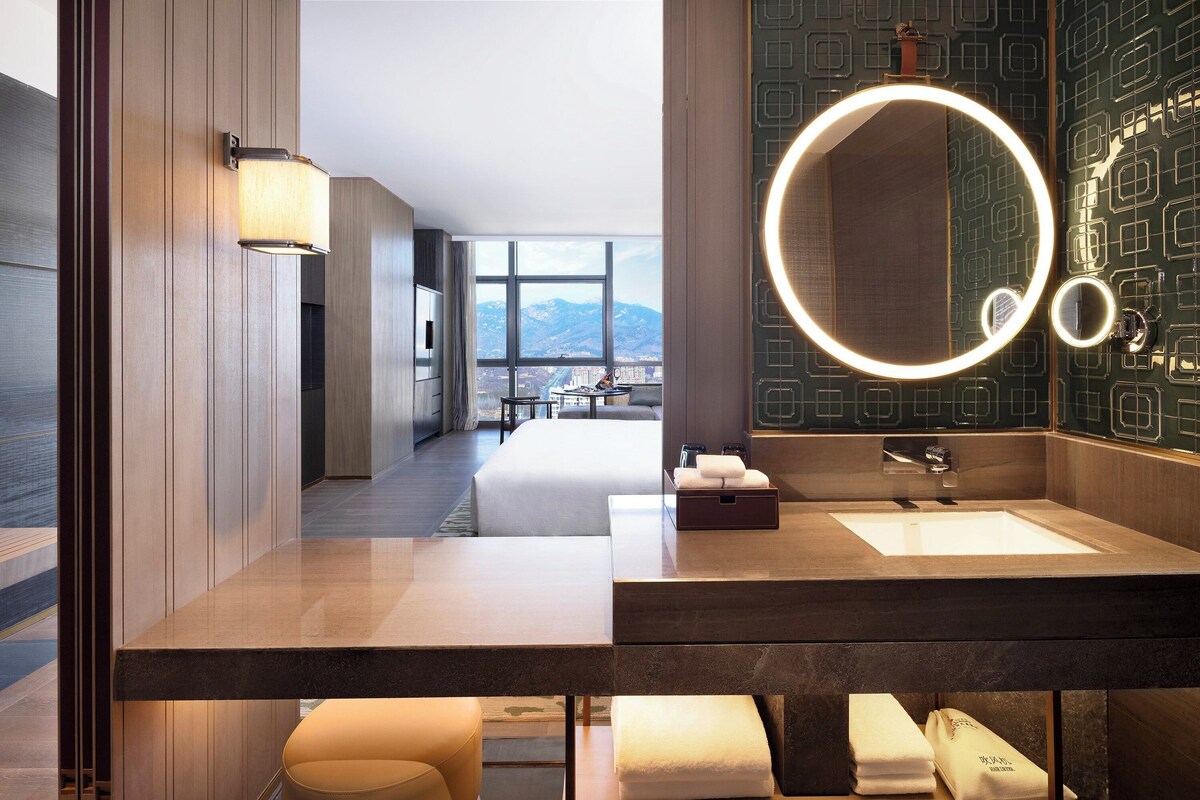 A modern hotel bathroom with a large mirror, a sink, and a bench.