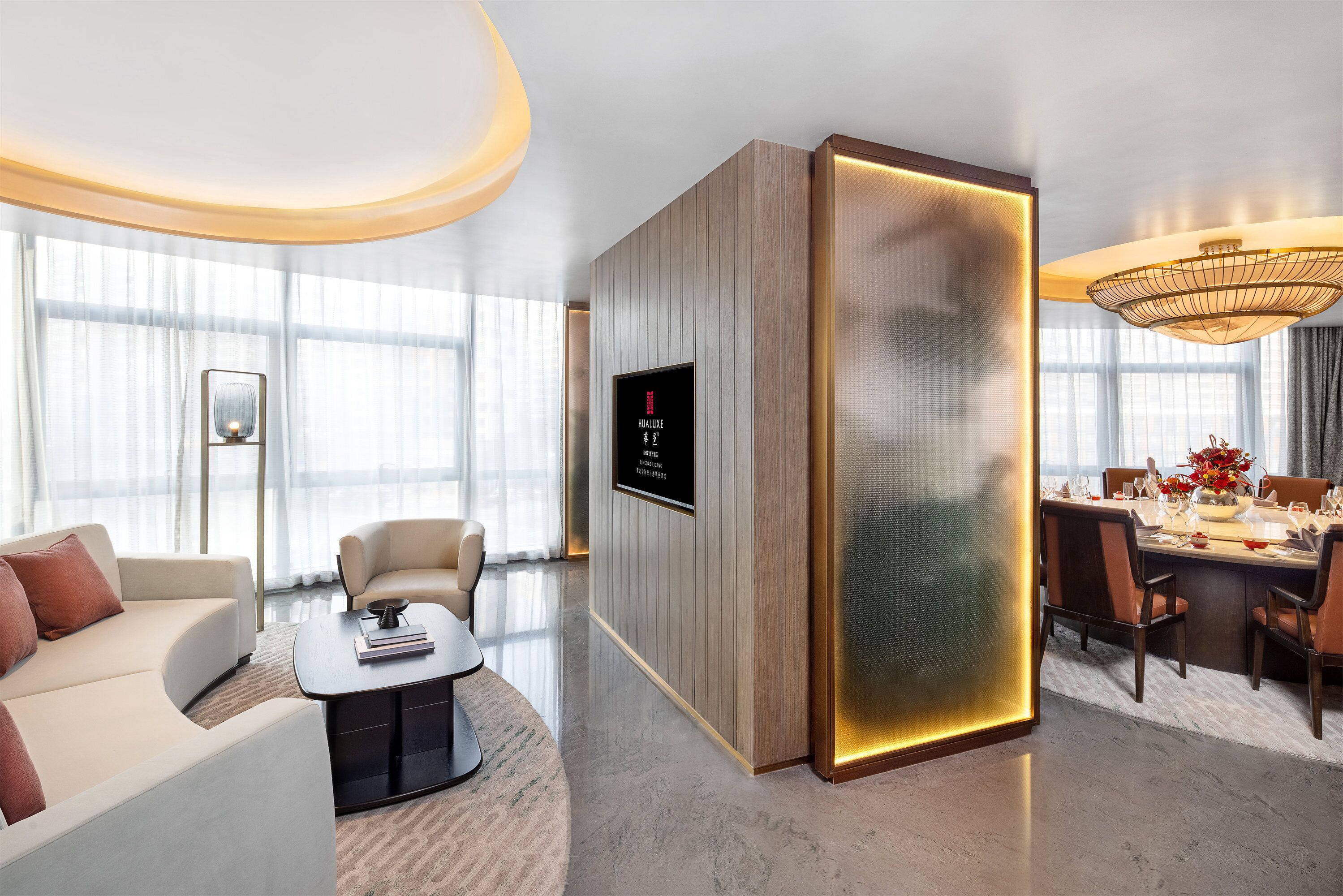 Een moderne hotellobby met een gebogen plafond, een zithoek met een bank en stoelen, een eetgedeelte met een tafel voor vier personen, en een grote tv aan de muur.