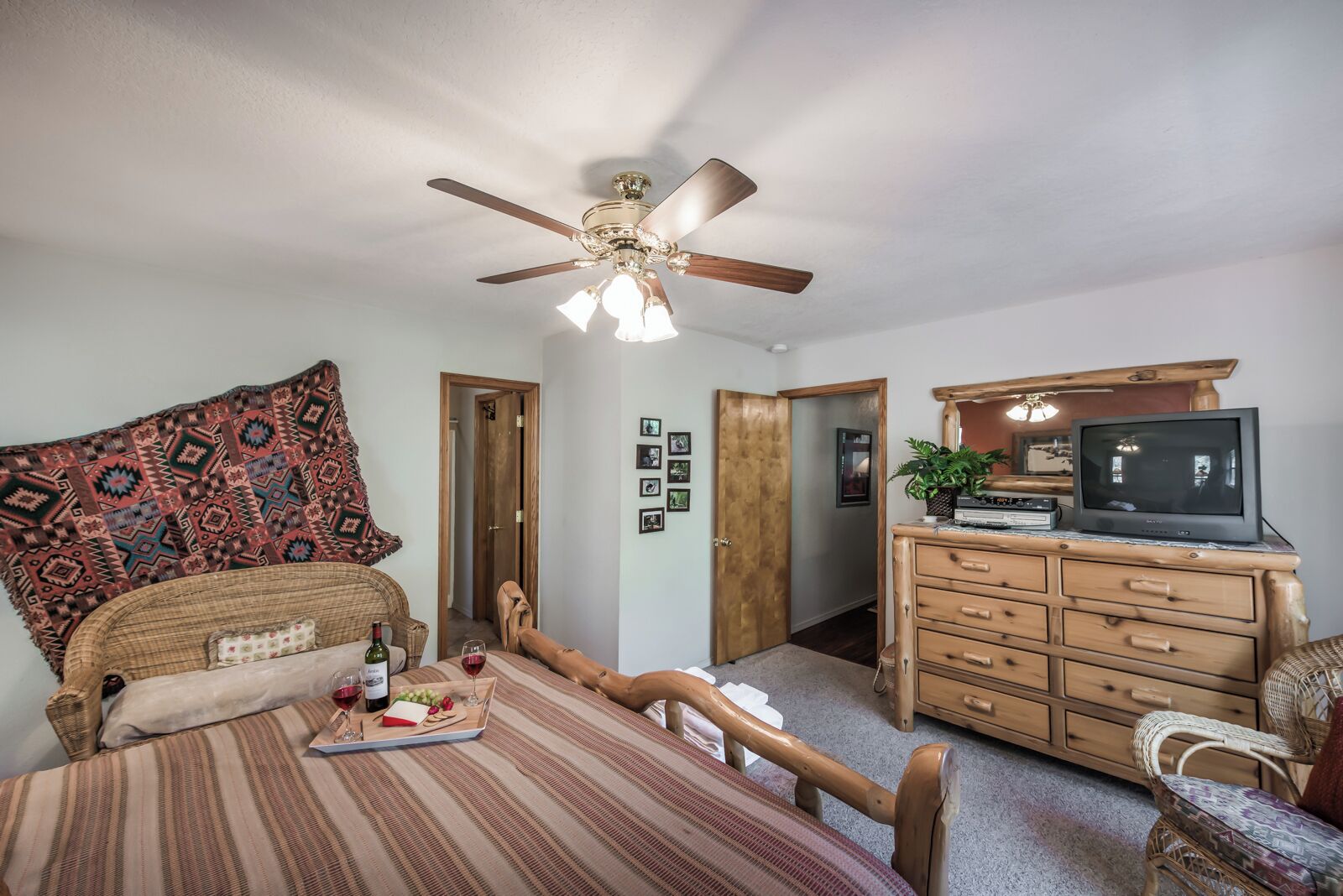 A cozy room with a bed, a dining table with wine glasses, a television on a wooden dresser, and a ceiling fan.