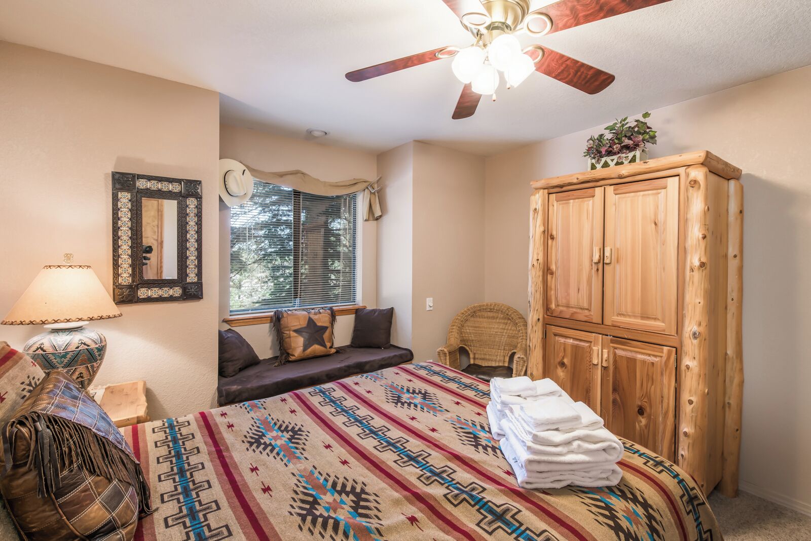A bedroom with a bed, a wooden wardrobe, a window with blinds, a ceiling fan, and a wall-mounted mirror.