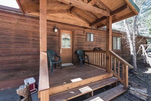 A wooden cabin with a porch, two chairs, a red cooler, and a bear sculpture.