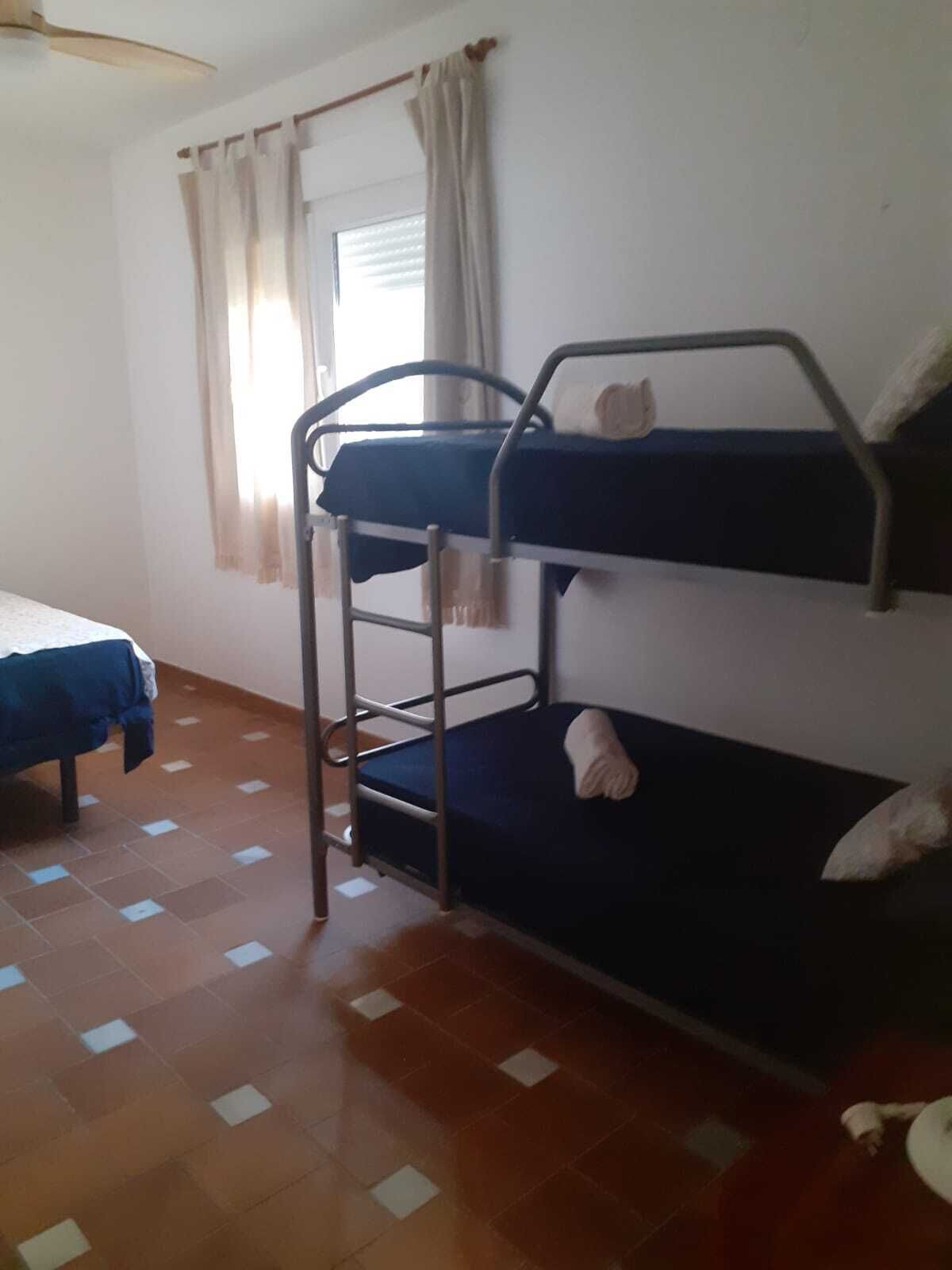 Une chambre de dortoir avec deux lits superposés, un lit simple, une fenêtre avec des rideaux et un sol carrelé.