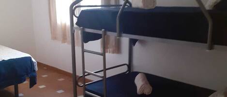 Une chambre de dortoir avec deux lits superposés, un lit simple, une fenêtre avec des rideaux et un sol carrelé.