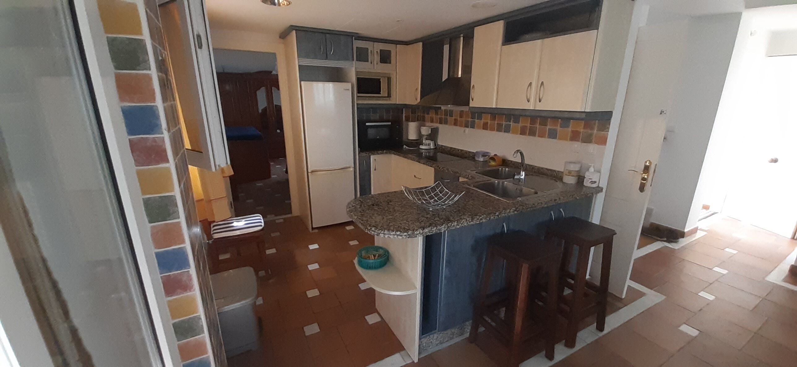 A compact kitchen with a refrigerator, microwave, and sink. There are two wooden stools and a small table. The floor has a checkered pattern.