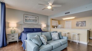 Une chambre avec un lit, un canapé bleu, un ventilateur de plafond et un coin cuisine équipé d’un comptoir et de tabourets.