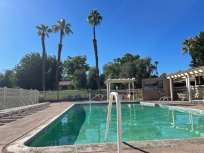 A clear swimming pool with a white pool ladder, surrounded by a white fence, palm trees, and a covered seating area. - Palm Desert airy two bedroom-steps from the pool!-golf, tennis, pickle ball!!! (Palm Desert)