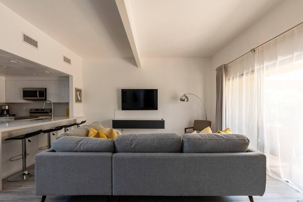 Ein modernes Wohnzimmer mit grauem Sofa, einem flachbildfernseher an der Wand und einer Küchenzeile mit Mikrowelle und Kaffeemaschine.