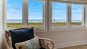 Una habitación con vistas a la playa a través de amplias ventanas, una silla de mimbre con cojín y suelo de madera.