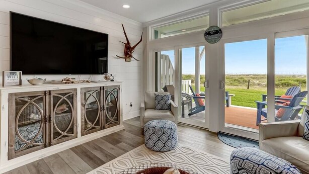 Un salón amplio con un televisor grande, una vitrina con puertas de cristal, otomanas estampadas y vistas al exterior a través de grandes puertas correderas.