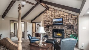 A living room with a stone fireplace, a television mounted above it, and a ceiling fan. - White Mountain Chateau by Ruidoso Vacation Rentals (Ruidoso)