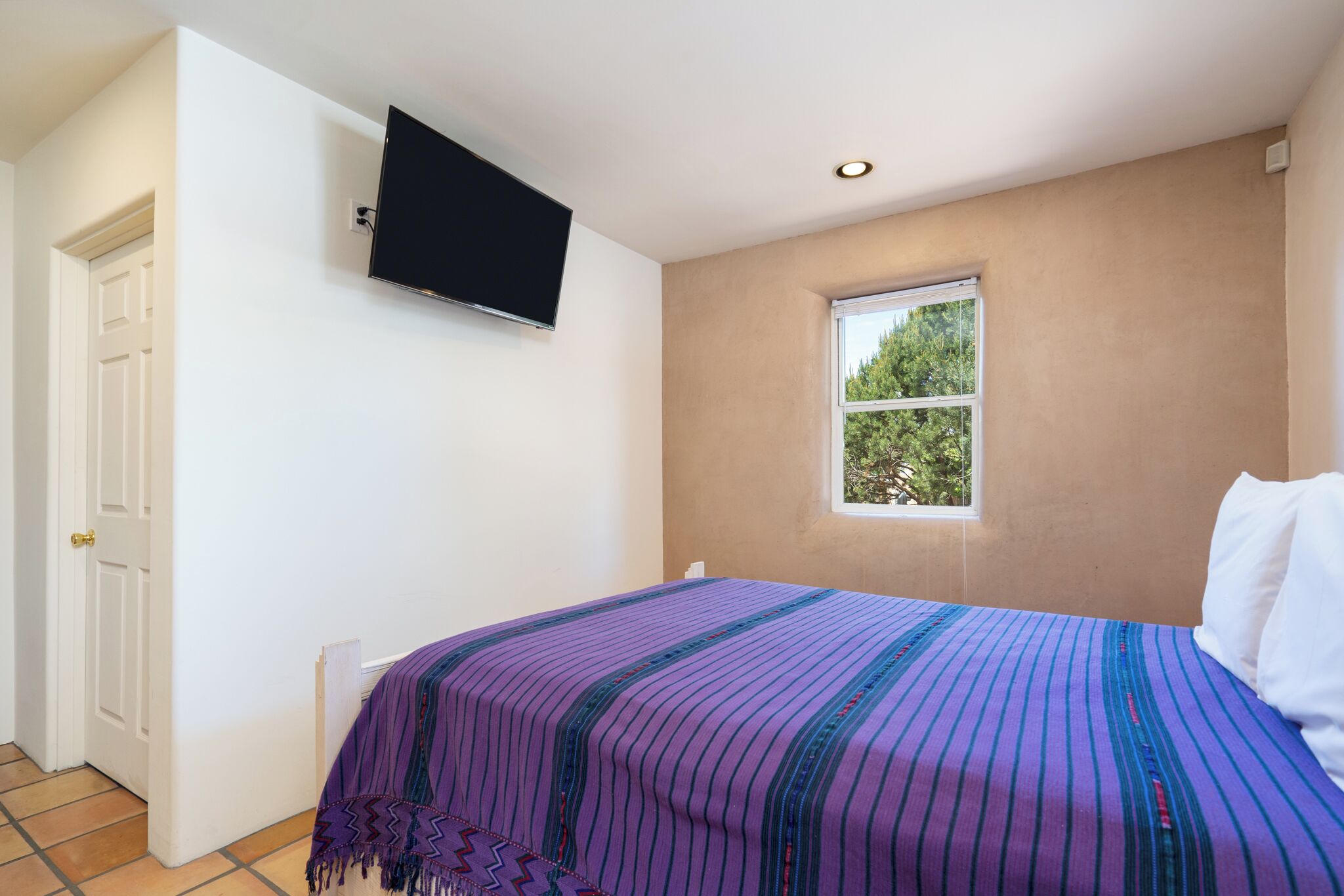 A bedroom with a bed, a television mounted on the wall, a window, and a door.