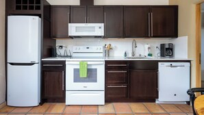A compact kitchen with dark wooden cabinets, a white refrigerator, a microwave, an oven, a dishwasher, and a coffee maker. - Casa Columbia by Kokopelli Property Management (Santa Fe)